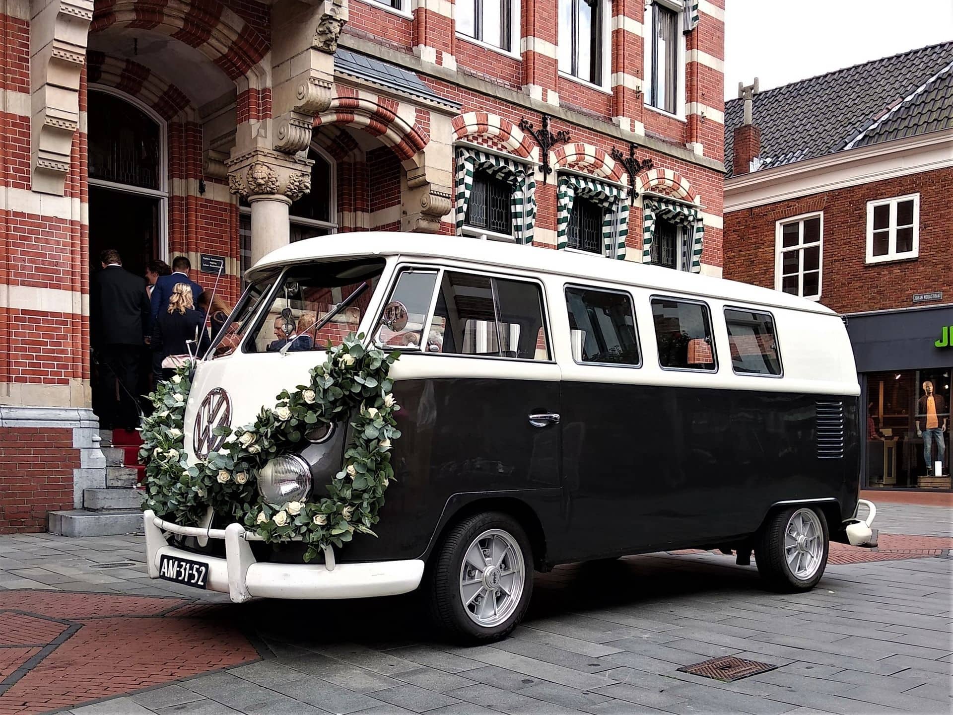 Schwarz-weißer VW Bulli mit Blumenkranz vor einem Backsteingebäude